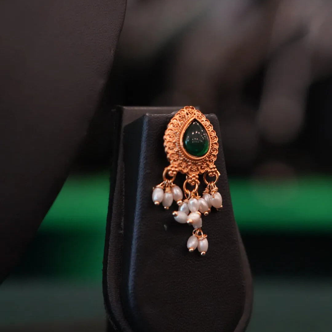 Ornate earring with green gemstone and pearl drops on a black stand.