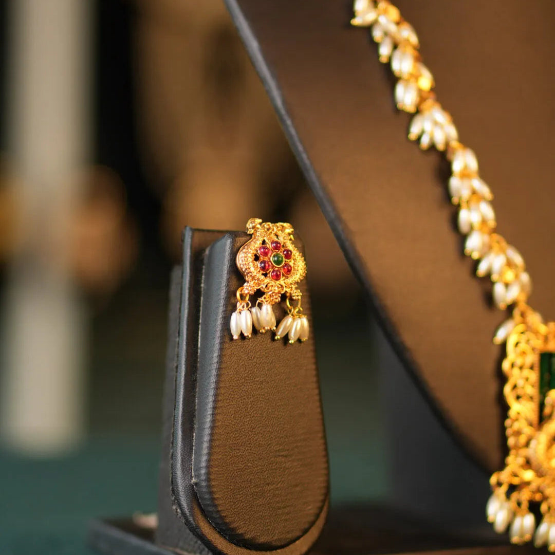 Gold earring with red gemstone on a brown stand, pearl necklace in the background